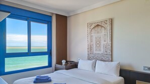Une chambre d’hôtel avec un grand lit, une table de chevet, une lampe de chevet, une œuvre d’art accrochée au mur et une vue sur l’océan.