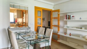 Un espace repas moderne avec une table et des chaises en verre, une cuisine en arrière-plan et une bibliothèque contenant des livres et des objets décoratifs.