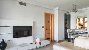Un salon moderne doté d’un téléviseur à écran plat, d’une table basse en verre ornée d’un vase, d’un canapé et d’un coin repas avec une table et des chaises en verre.