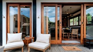 Un terrazzo in legno con due poltrone bianche imbottite e un tavolino, una porta scorrevole in vetro che dà su un'area pranzo con tavolo e sedie, e vista sul paesaggio circostante.