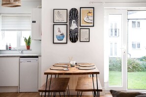 A modern kitchen with a dining table set for four, a dishwasher, and a window with a view of a garden. - Stylish Apartment, Garden with Beautiful Sea Views (Newquay)