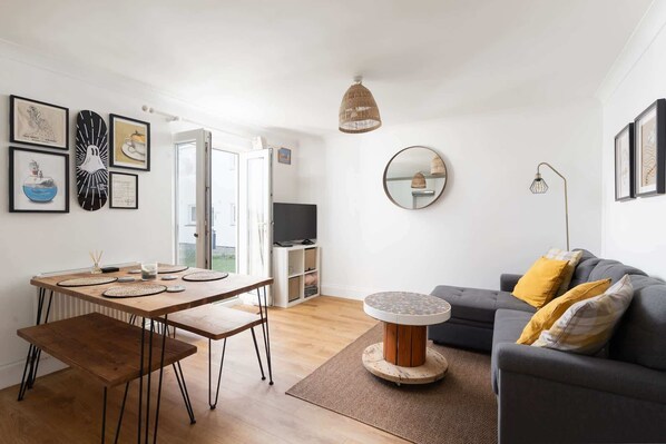 A modern living room with a dining area, a grey sofa with yellow cushions, a round coffee table, and a TV in the background. - Stylish Apartment, Garden with Beautiful Sea Views (Newquay)