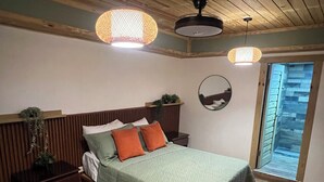 Un dormitorio con techo de madera, cama con ropa de cama verde y almohadas naranjas, una mesita de noche y una ventana con vista a un muro de piedra.