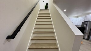 Un escalier avec des marches en bois et une rampe noire, menant à un espace cuisine doté de meubles blancs et d’un réfrigérateur en acier inoxydable.
