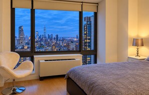 A hotel room with a large window, a bed, a chair, a bedside table with a lamp, and a wall-mounted heater. - Manhattan Lux High-Rise next to Empire State Bldg (New York)