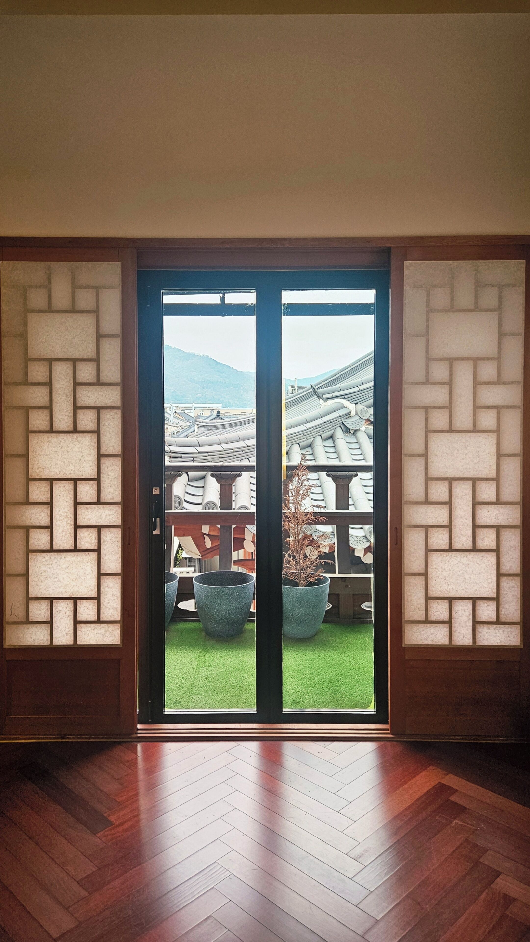A room with a herringbone wood floor, a sliding glass door leading to a balcony with a view of traditional tiled roofs and a grassy area.