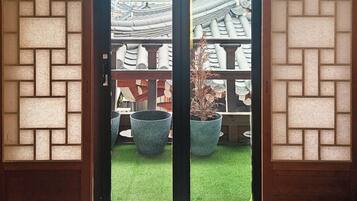 A room with a herringbone wood floor, a sliding glass door leading to a balcony with a view of traditional tiled roofs and a grassy area.
