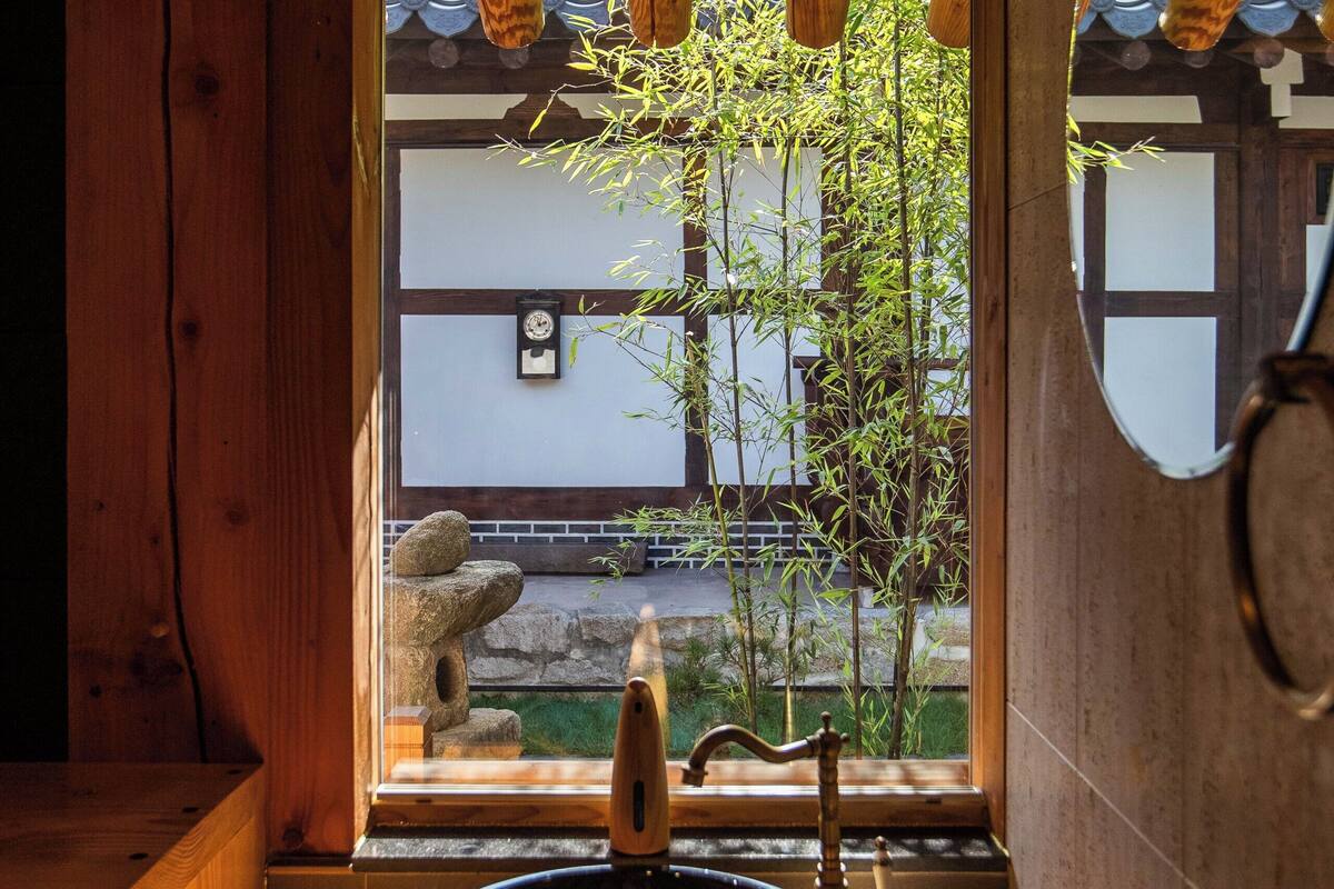 A wooden interior with a sink, a mirror, and a view of bamboo outside.