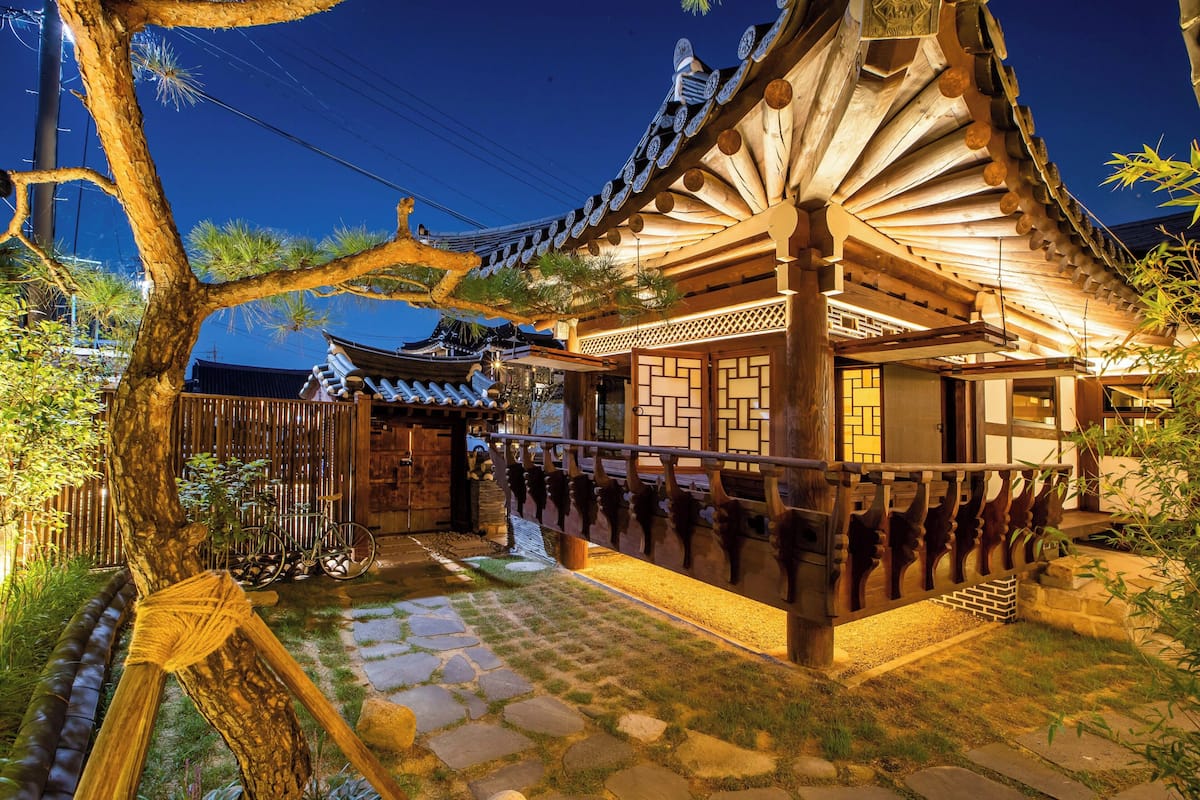 A traditional Japanese building with a wooden structure, stone pathway, and a tree in the foreground.