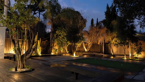 A well-lit outdoor patio area with a pool, benches, and various trees. - Palo Alto Villa (East Palo Alto)
