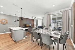 Una cocina moderna con una isla central, comedor con sillas grises, televisor de pantalla plana y ventilador de techo.