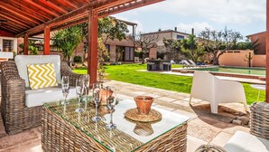 Un patio al aire libre con mobiliario de mimbre, una mesa con cubierta de vidrio y vistas a una piscina y jardín.