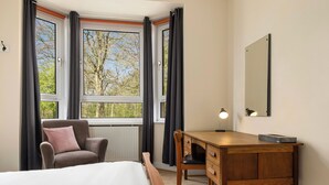 Ein Schlafzimmer mit Bett, Sessel, Schreibtisch, einem Fenster mit Vorhängen und einer Heizung.