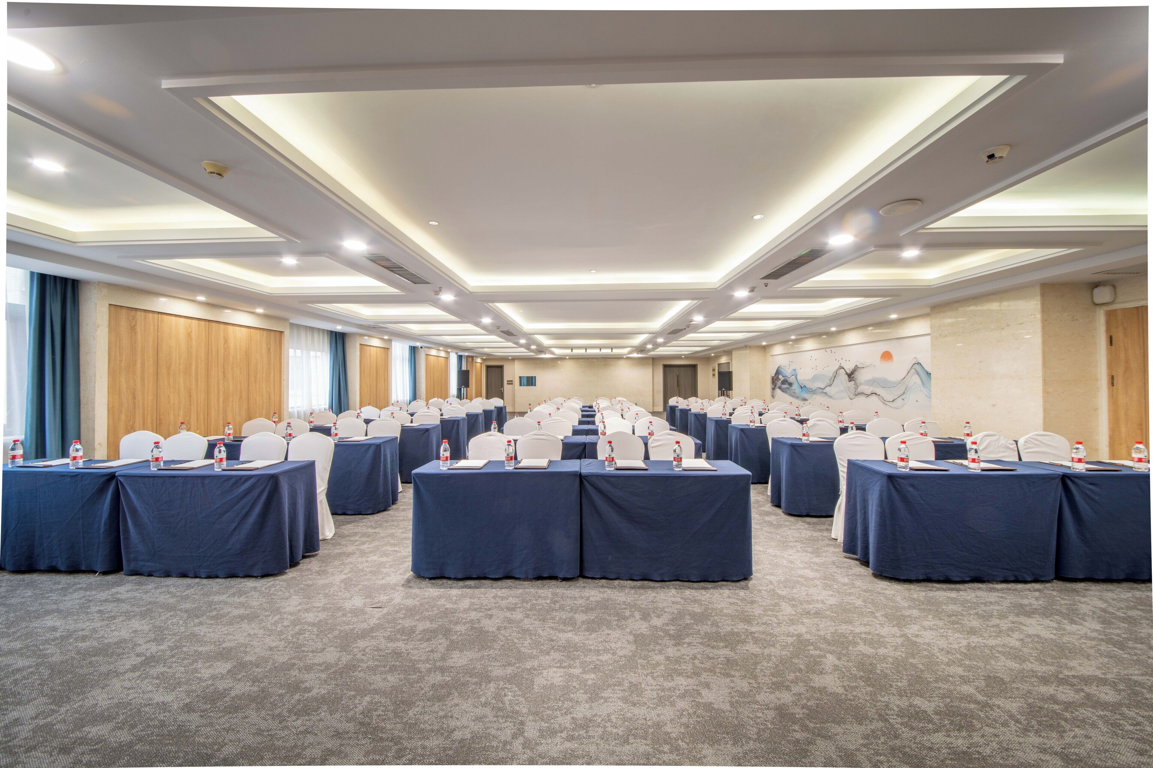A conference room with multiple tables, each with a blue tablecloth and white chairs. The room has a carpeted floor and a ceiling with recessed lighting.