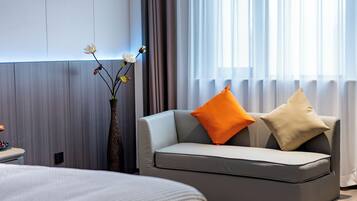 Habitación de hotel moderna con un sofá gris, dos almohadas, un jarrón con flores y una cama con cubrecama blanca.