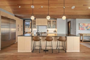 A modern kitchen with a central island, bar stools, pendant lighting, and a stainless steel refrigerator.