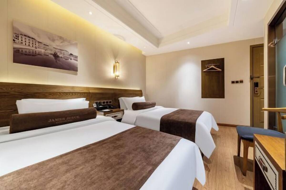 A hotel room with two beds, a desk, a chair, a coat rack, and a wall-mounted light fixture.