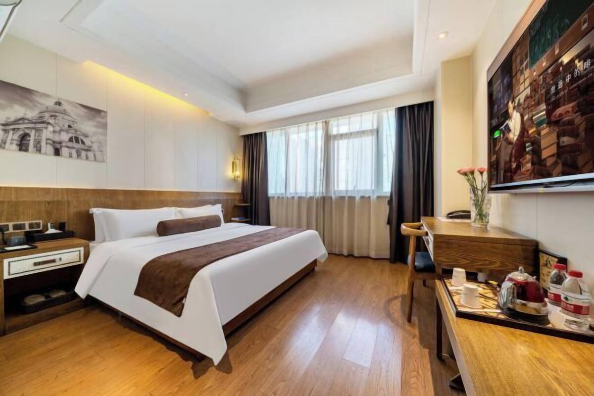 A hotel room with a large bed, wooden flooring, a desk with a tray of items, a painting on the wall, and a window with curtains.