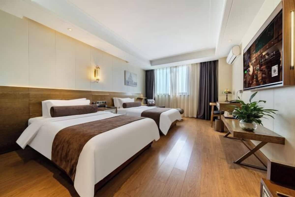A hotel room with two beds, a wooden floor, a desk with a plant, and a large window with curtains.