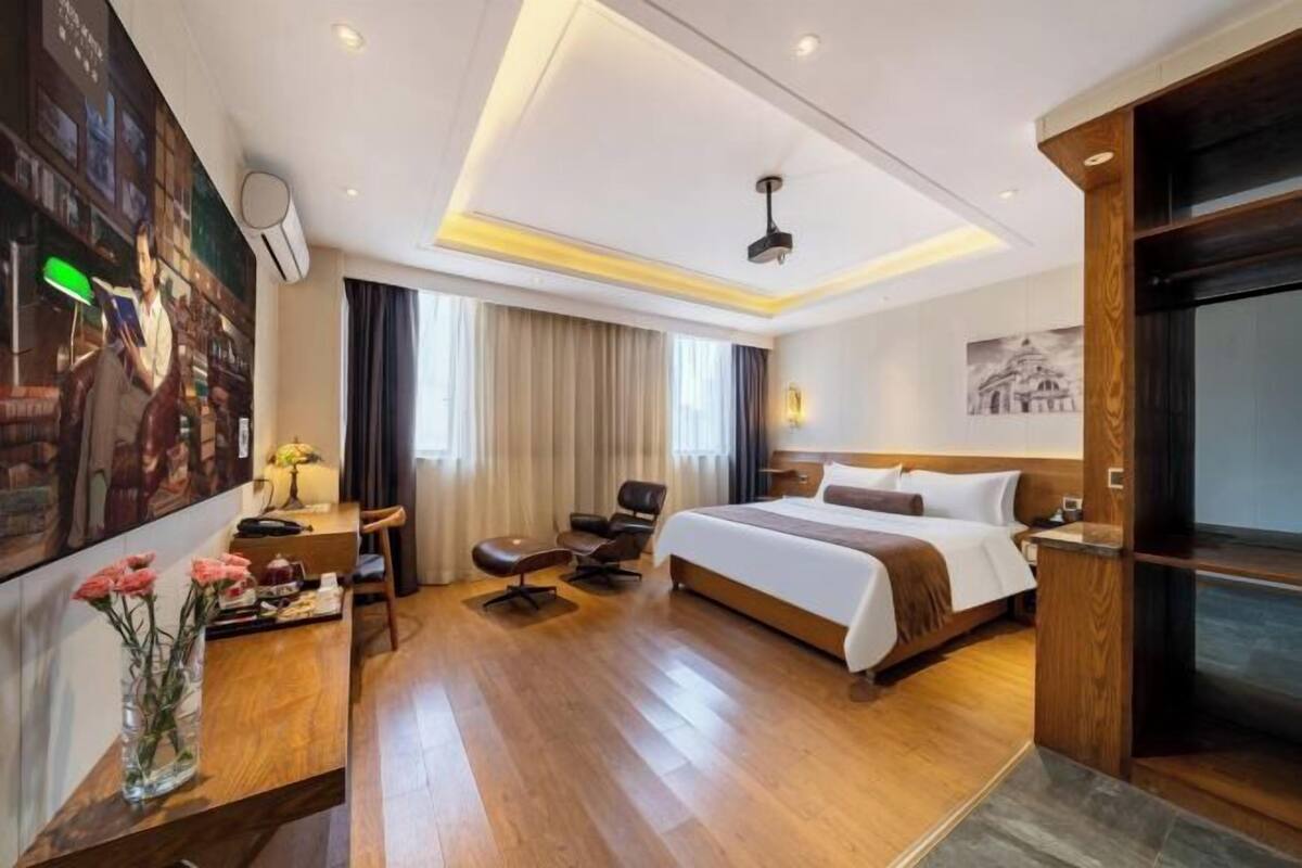 A hotel room with a large bed, a desk with a chair, a small table with flowers, and a large window with curtains.
