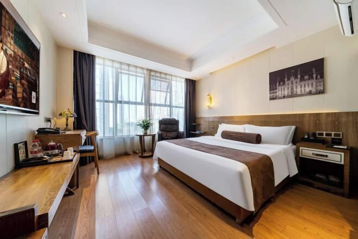 A hotel room with a large bed, a desk with a chair, a small table with a plant, and a large window with curtains.