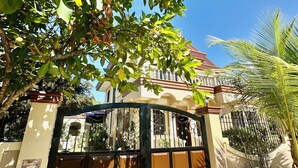 A gated entrance with a tiled floor, a metal gate, and a building with a tiled roof and windows in the background. - Spacious 3-bedroom house in Subic with AC, WiFi, Pool & Parking. Relax & unwind (Subic)