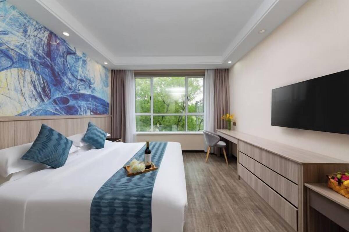 Quarto de hotel com cama grande, escrivaninha, televisão e janela com vista para árvores.