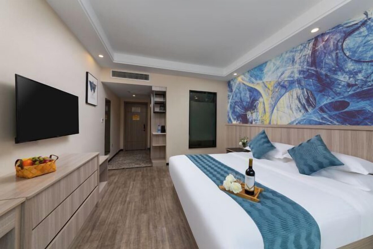 A modern hotel room with a large bed, a flat-screen TV, a fruit basket, and a painting on the wall.