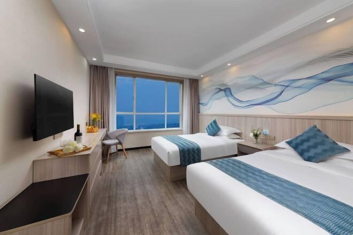 Quarto de hotel com duas camas, televisão, secretária e uma janela grande com vista.