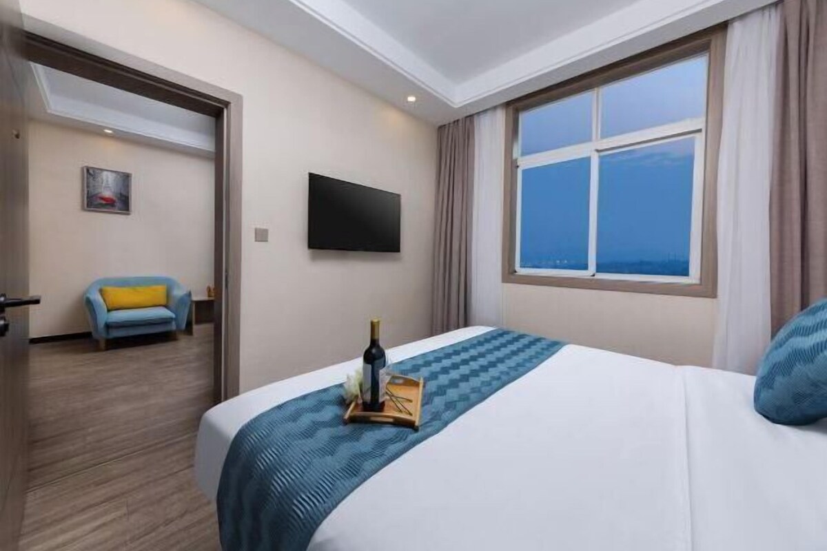 A hotel room with a bed, a flat-screen TV, a window with a city view, and a chair with a yellow cushion.