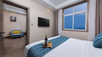 A hotel room with a bed, a flat-screen TV, a window with a city view, and a chair with a yellow cushion.