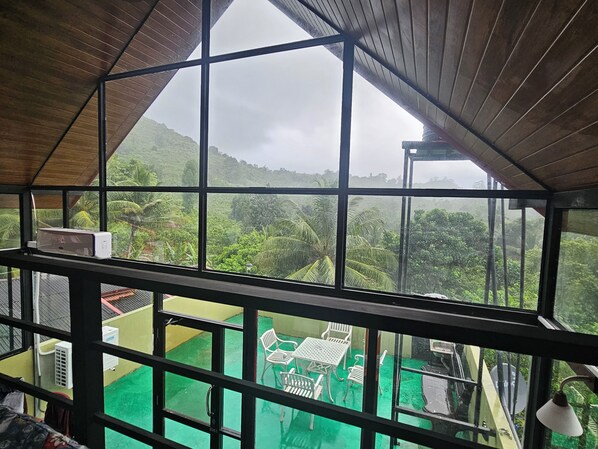 A room with a large window overlooking a lush green landscape. Inside, there is a green floor, a white table with chairs, and a white air conditioning unit.