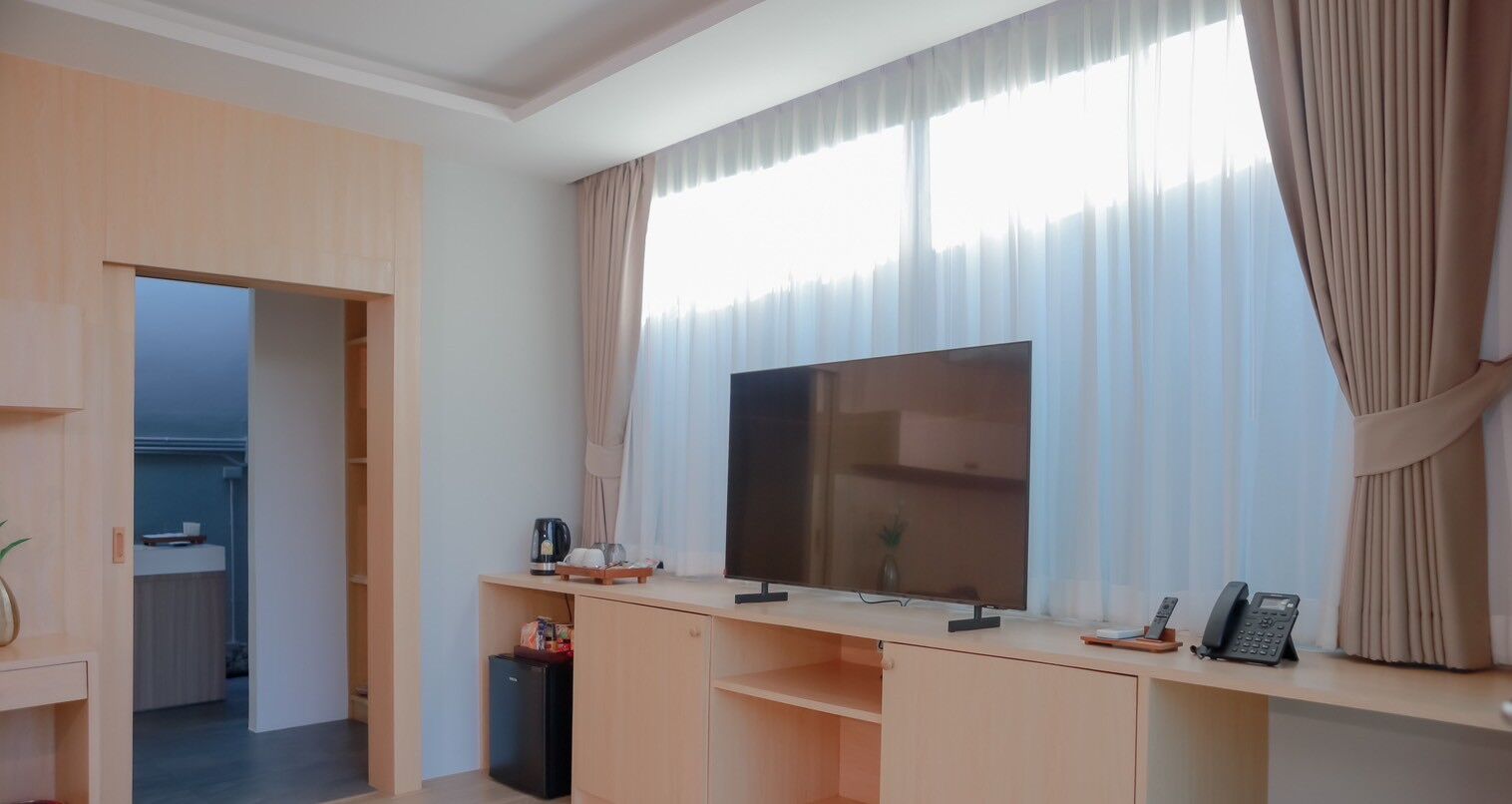 Une chambre d’hôtel moderne dotée d’une grande télévision à écran plat, d’un bureau avec un téléphone et une petite table, ainsi que d’une fenêtre avec des rideaux légers.