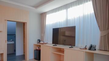 Une chambre d’hôtel moderne dotée d’une grande télévision à écran plat, d’un bureau avec un téléphone et une petite table, ainsi que d’une fenêtre avec des rideaux légers.