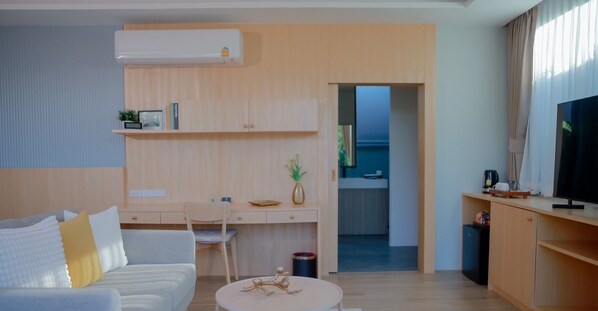 Una sala moderna con sofá, una mesa de centro, televisor y un escritorio con silla. Se observa una puerta que da a una zona de cocina.