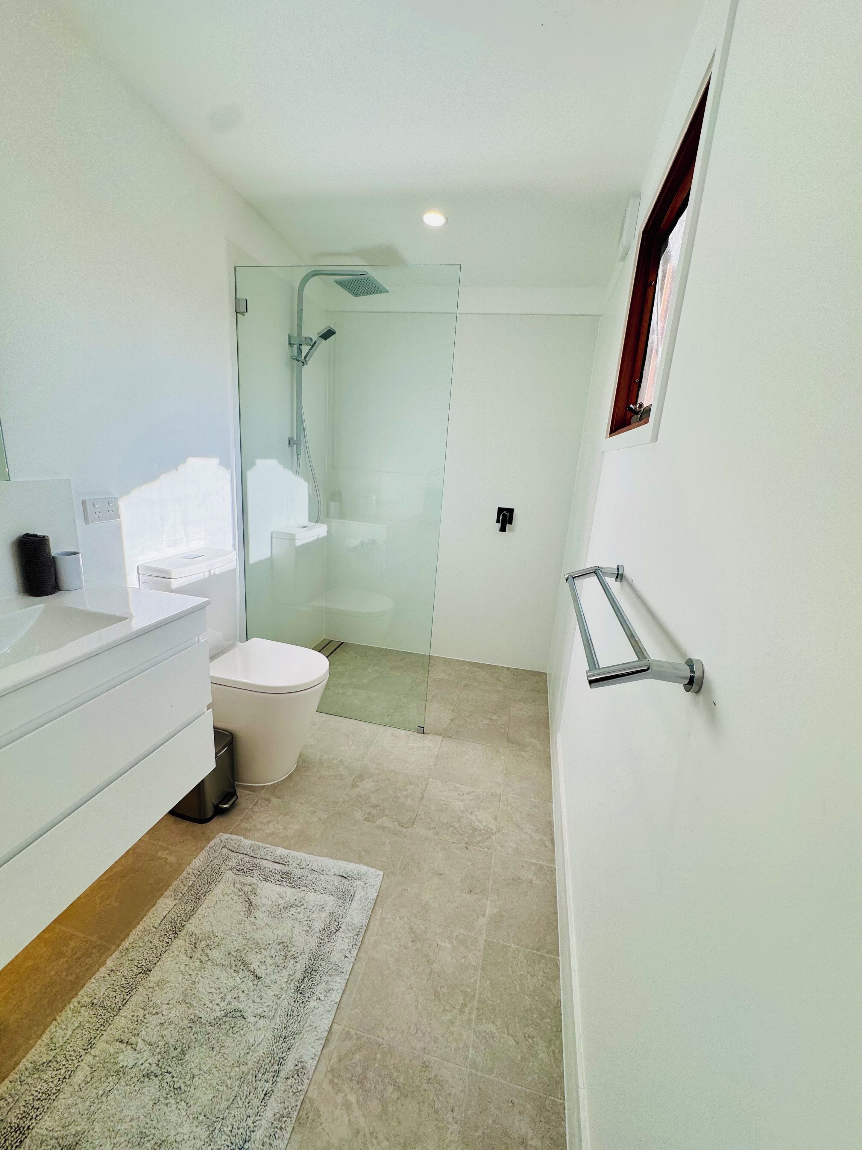 A modern bathroom with a glass shower enclosure, a toilet, a sink with a towel holder, and a small window.