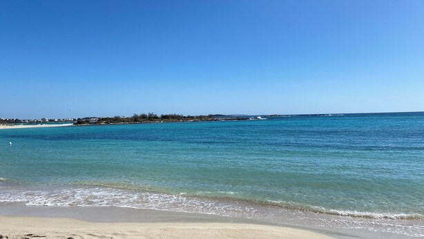 A beach with clear blue water, a sandy shore, and a distant shoreline with buildings and vegetation.