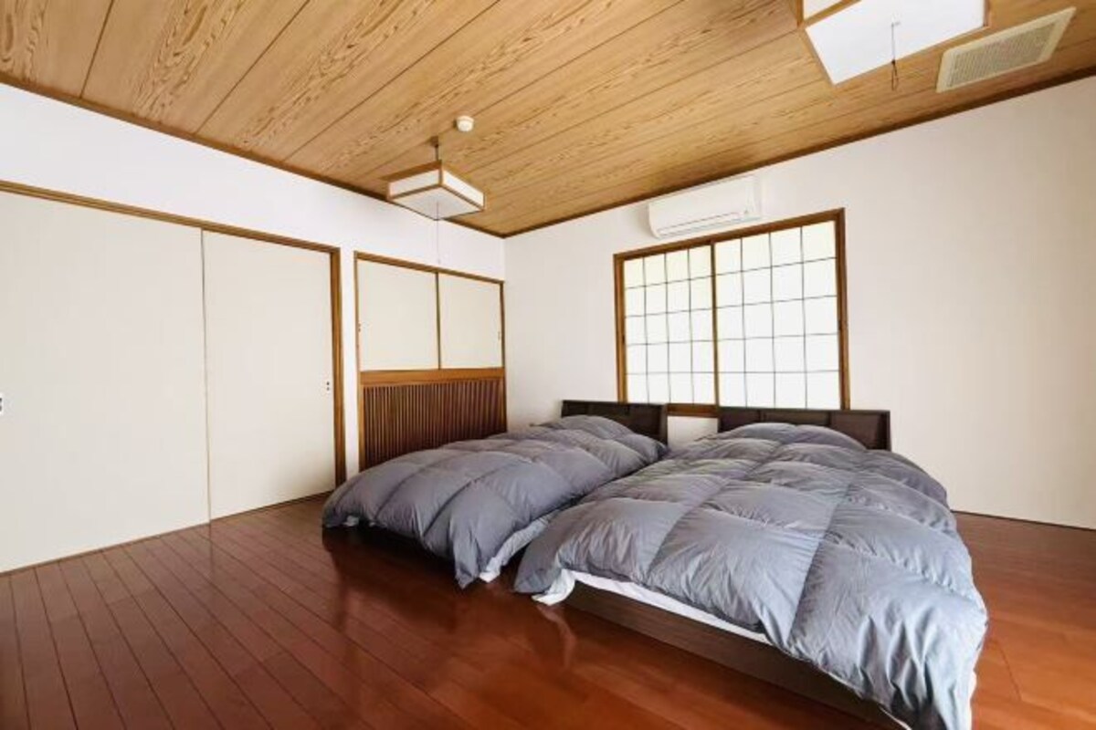 Ein Schlafzimmer mit Holzboden, einem großen Bett mit grauer Bettwäsche, einem Holz-Kopfteil und einem Fenster mit Gittermuster.