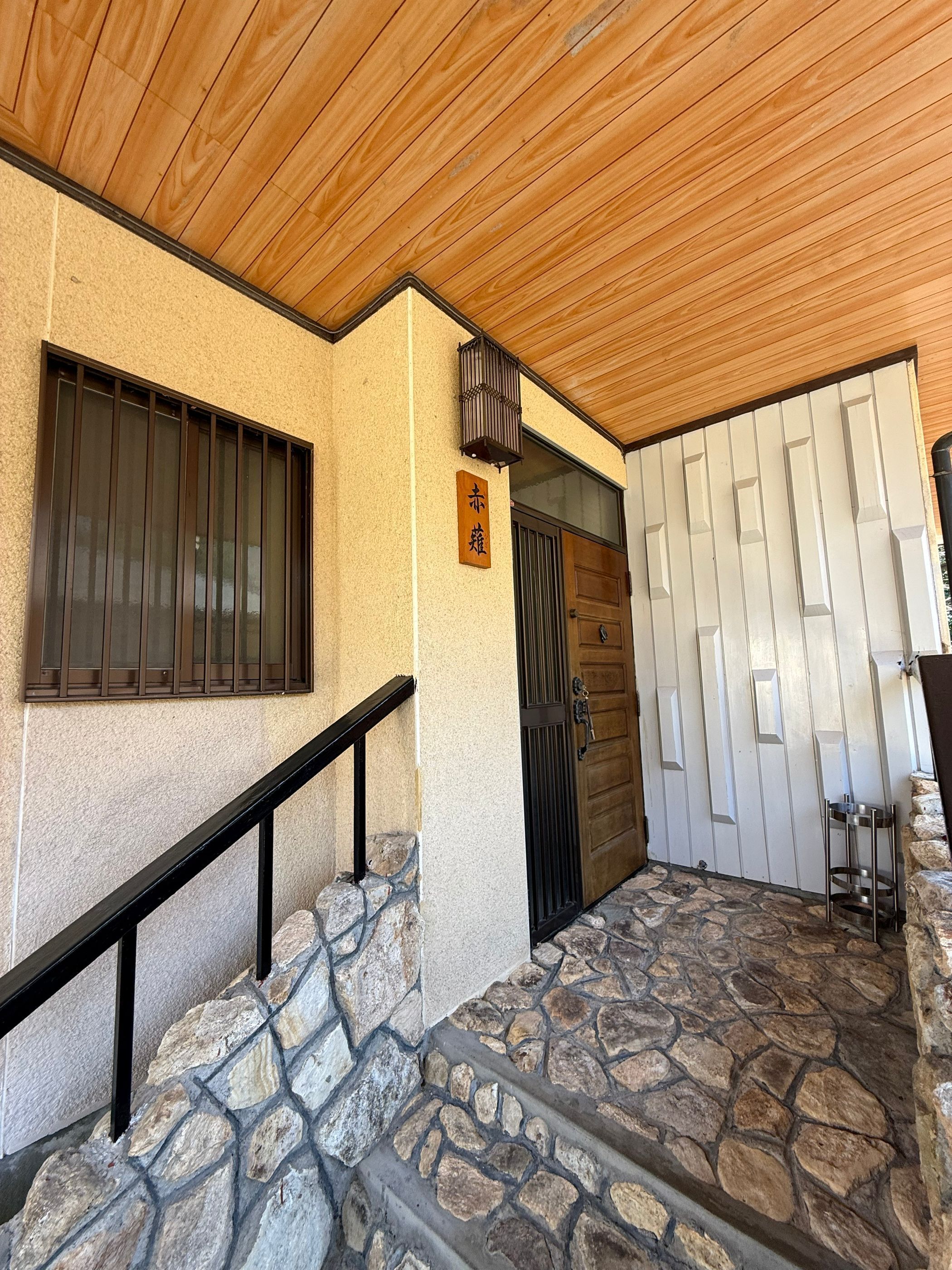 Una escalera de piedra que conduce a una puerta de madera con una reja de metal, una ventana con persianas verticales y un farol en la pared.