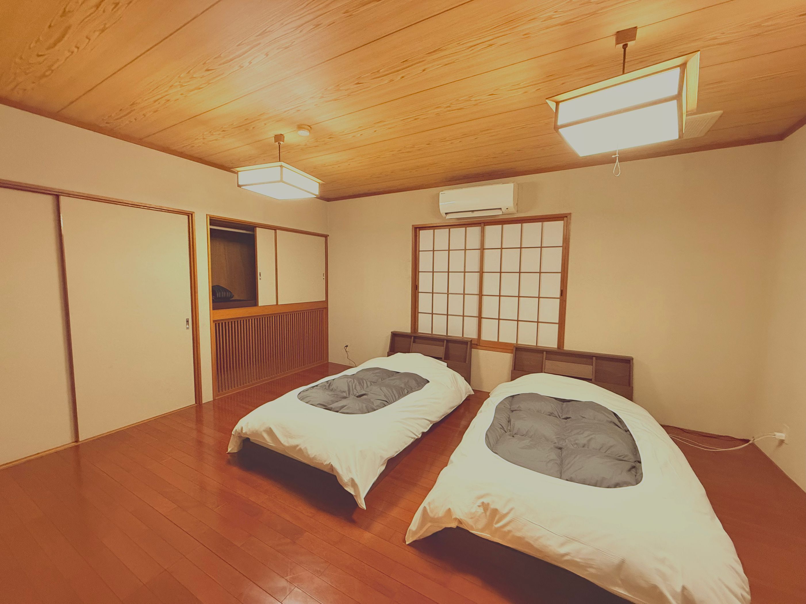 A room with two beds, wooden flooring, a sliding door, a window with a grid pattern, and a ceiling light fixture.