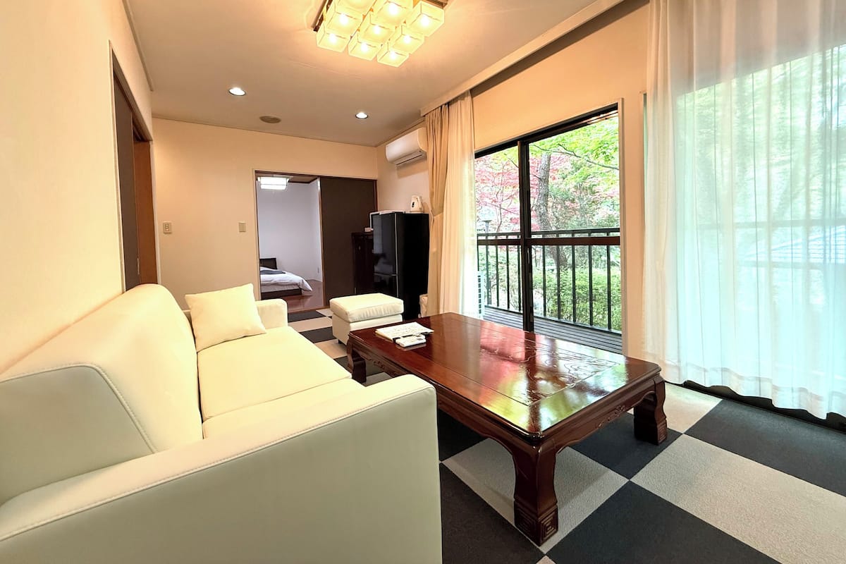 Habitación de hotel con sofá, una mesita de madera, televisor y ventana grande con vistas a árboles.