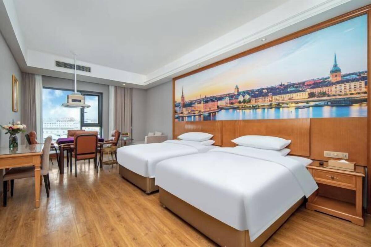 Hotellihuone, jossa on kaksi sänkyä, työpöytä, tuoleja, suuri ikkuna ja kaupunkimaisemaa esittävä taulu.
