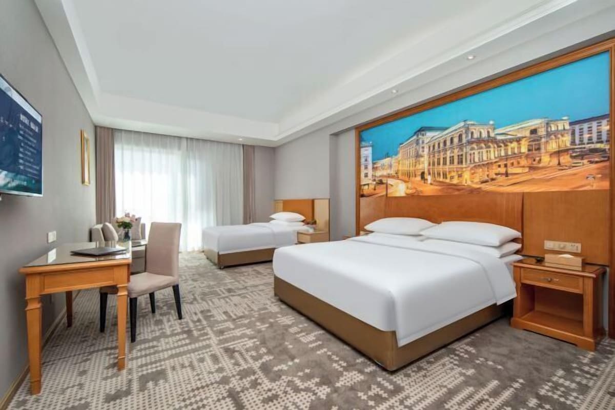 A hotel room with two beds, a desk, a chair, and a large painting on the wall.