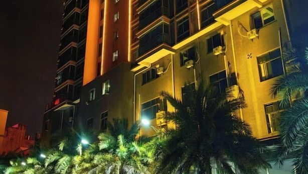 A multi-story building with balconies and air conditioning units, surrounded by palm trees and illuminated by artificial lighting.