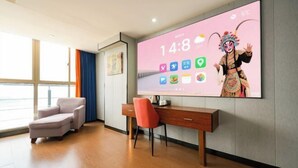 A modern hotel room with a large TV on the wall, a wooden desk, a comfortable chair, and a view through the window.