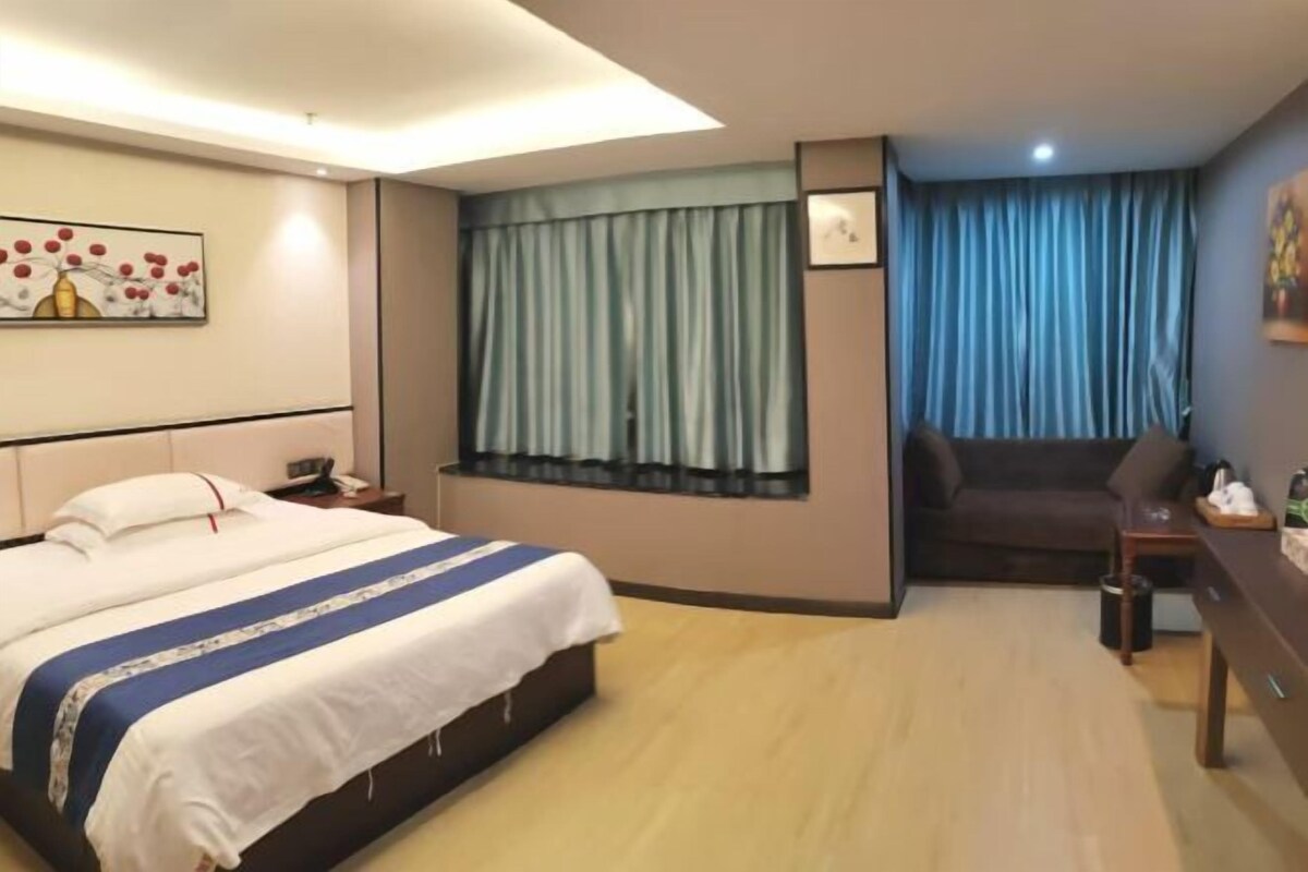 A hotel room with a bed, a window with curtains, a sofa, a desk, and a painting on the wall.