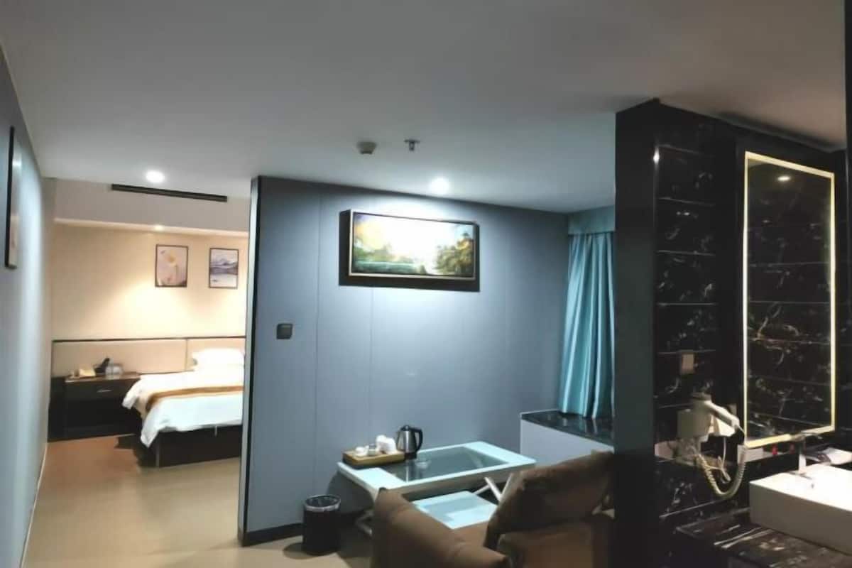 Habitación de hotel con cama, escritorio, silla, una mesita con hervidor de agua y baño con espejo y lavamanos.