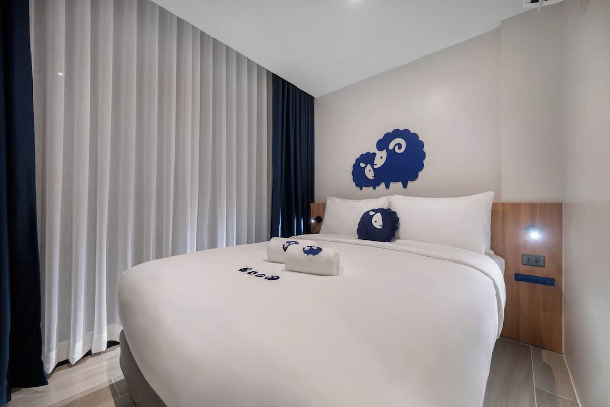 Une chambre d’hôtel moderne avec un grand lit, une décoration murale représentant un mouton, une tête de lit en bois et une table de chevet.
