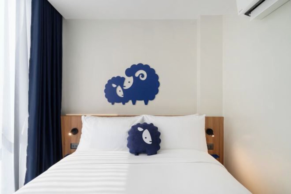 Une chambre d’hôtel avec un lit, deux oreillers, une œuvre d’art représentant des moutons accrochée au mur et une fenêtre avec des rideaux.