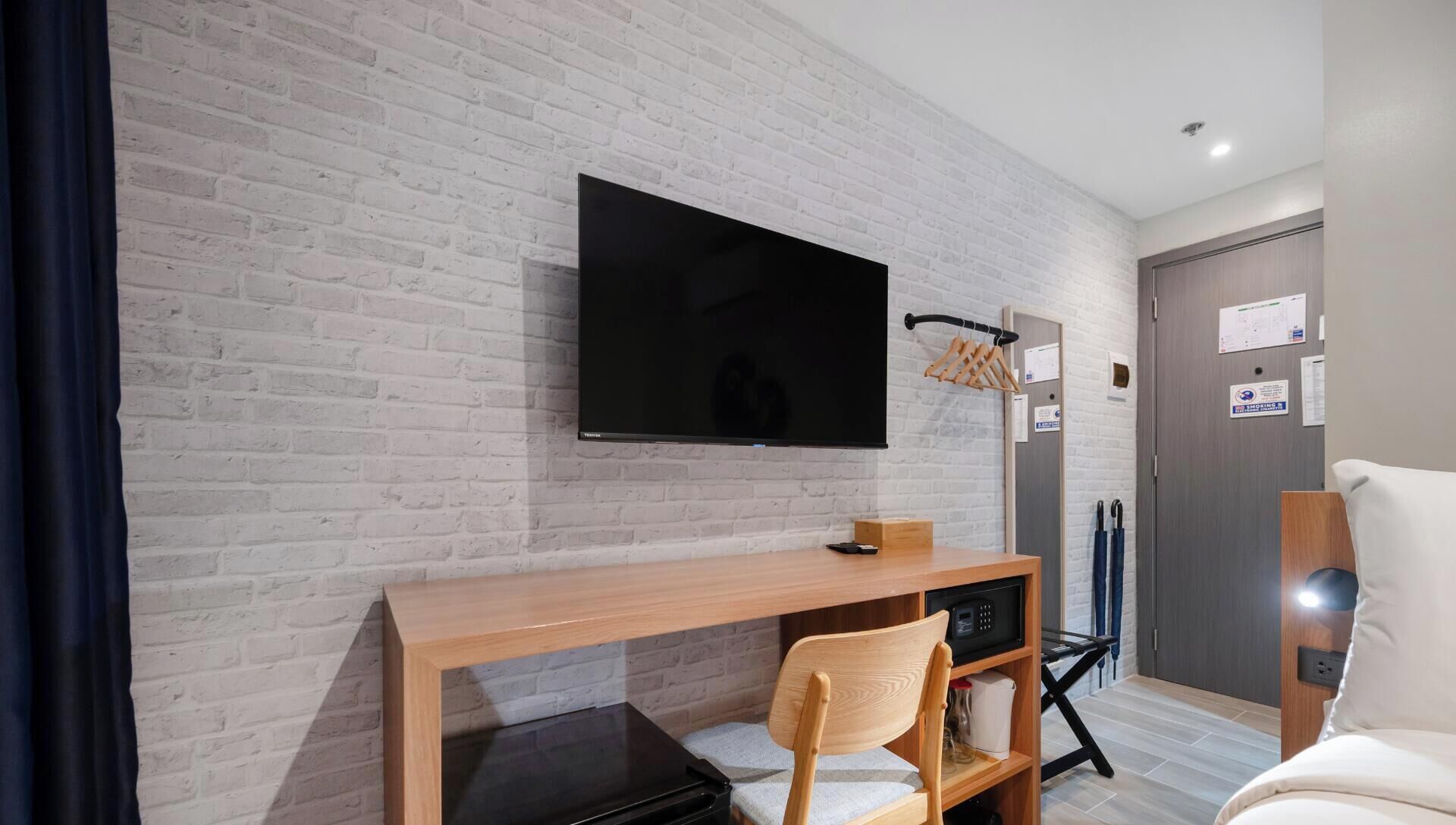 A hotel room with a brick wall, a flat-screen TV mounted above a wooden desk, a chair, and a bed.